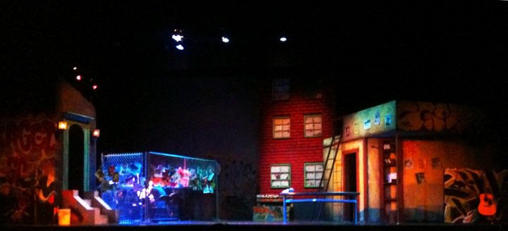 The RENT set: a hand-built two-story tenement, chain-link fence, and graffiti, lit in red and blue stage light.
