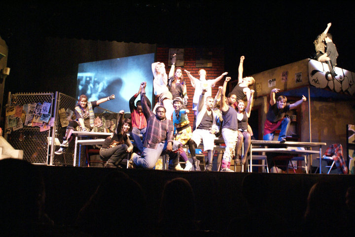 The full RENT cast on stage with raised fists, gathered around the tenement set.